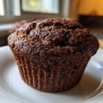 Close-up of a single, dark, moist bran muffin sitting on a white plate near a window.