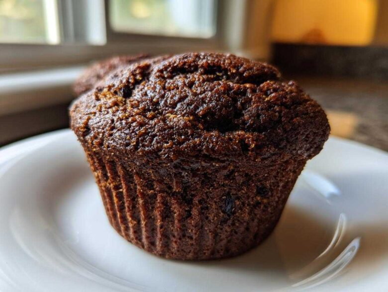 Close-up of a single, dark, moist bran muffin sitting on a white plate near a window.