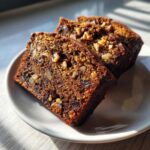 Three slices of rich, dark date nut bread packed with visible walnuts, served on a white plate.
