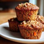 A stack of three moist morning glory muffins topped with chopped walnuts on a light-colored plate.