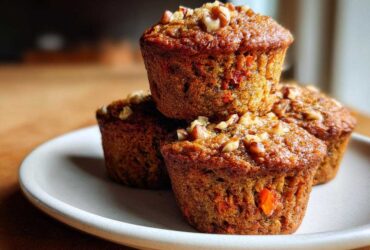 A stack of three moist morning glory muffins topped with chopped walnuts on a light-colored plate.