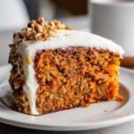 Close-up of a moist slice of carrot cake recipe, topped with thick cream cheese frosting and chopped walnuts.