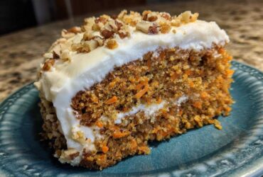 A close-up of a moist slice of carrot cake recipe topped with thick cream cheese frosting and chopped pecans.