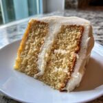 Close-up of a moist slice of vanilla cake recipe with thick white frosting layers.