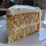 A close-up of a moist slice of vanilla cake recipe with white frosting between layers.