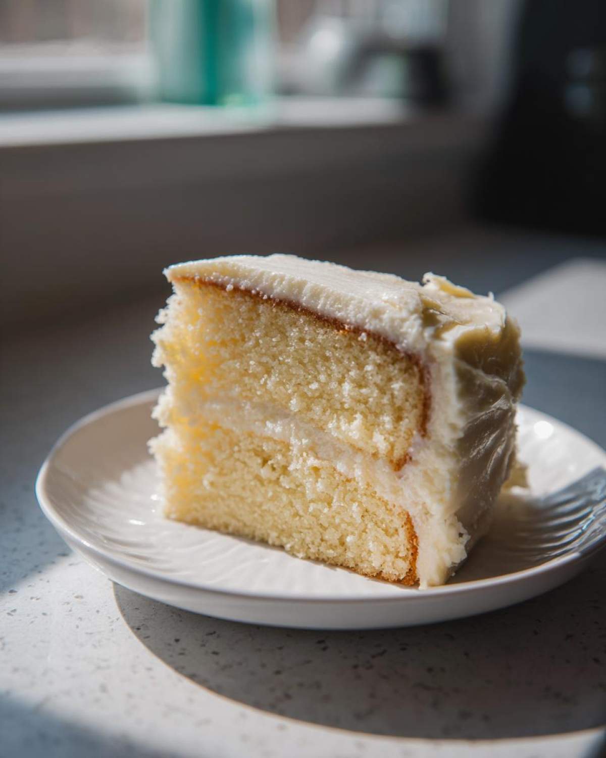 A moist slice of vanilla cake recipe with thick white frosting served on a small white plate.