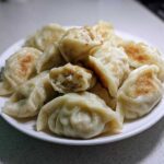 A white plate piled high with golden-brown pan-fried Traditional Chinese Dumplings, one is broken open showing the filling.