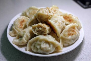 A white plate piled high with golden-brown pan-fried Traditional Chinese Dumplings, one is broken open showing the filling.