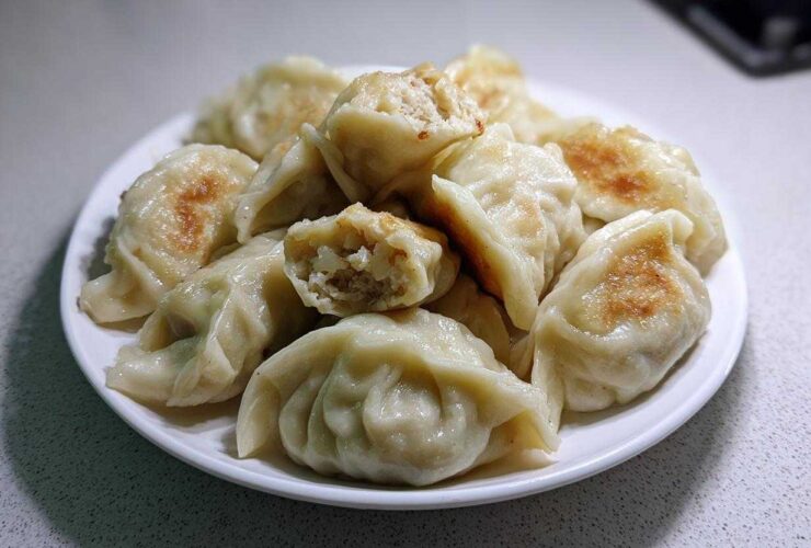A white plate piled high with golden-brown pan-fried Traditional Chinese Dumplings, one is broken open showing the filling.