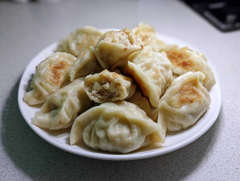 A white plate piled high with golden-brown pan-fried Traditional Chinese Dumplings, one is broken open showing the filling.