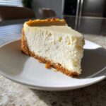 A perfect slice of creamy, baked Protein Cheesecake with a thick graham cracker crust served on a white plate.
