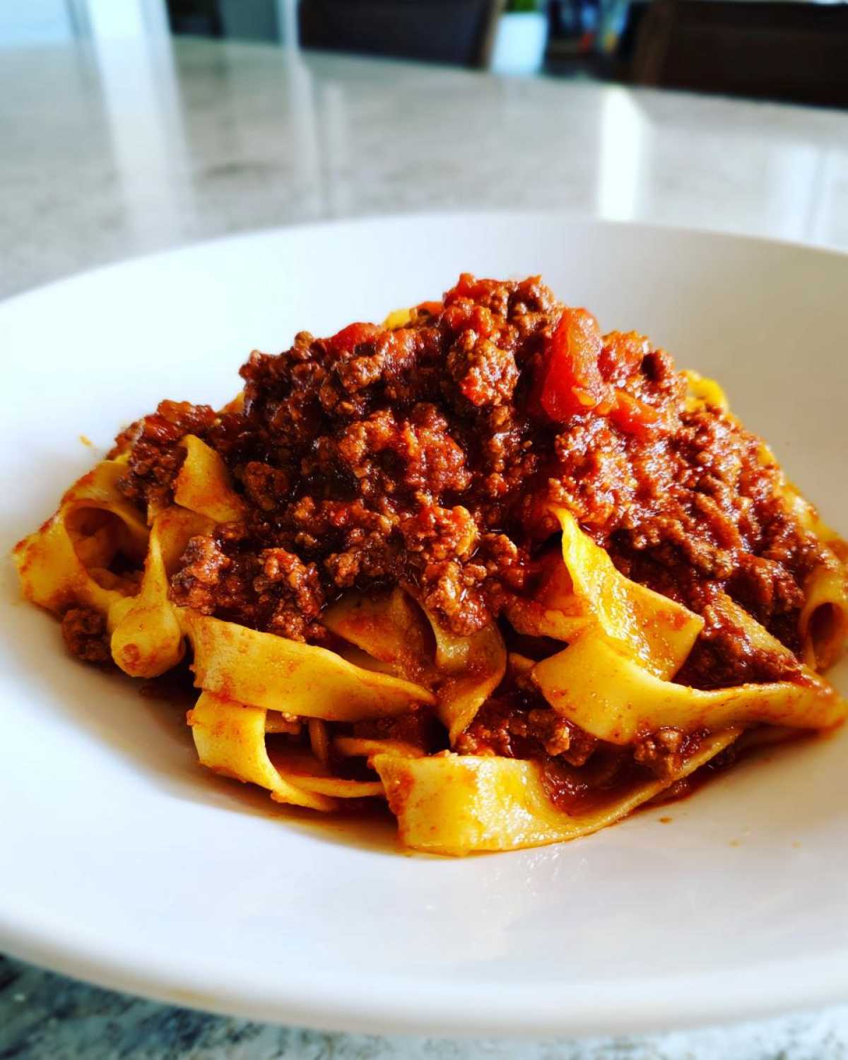 Close-up of rich bolognese sauce generously spooned over wide, fresh pappardelle pasta in a white bowl.
