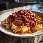 A close-up of rich bolognese sauce generously spooned over wide egg noodles on a white plate.