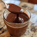 A spoon dripping thick, glossy chocolate sauce into a glass jar, perfect for chocolate overnight oats.
