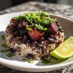 A beautifully plated serving of savory black beans and rice topped with sausage chunks and fresh cilantro, served with a lime wedge.