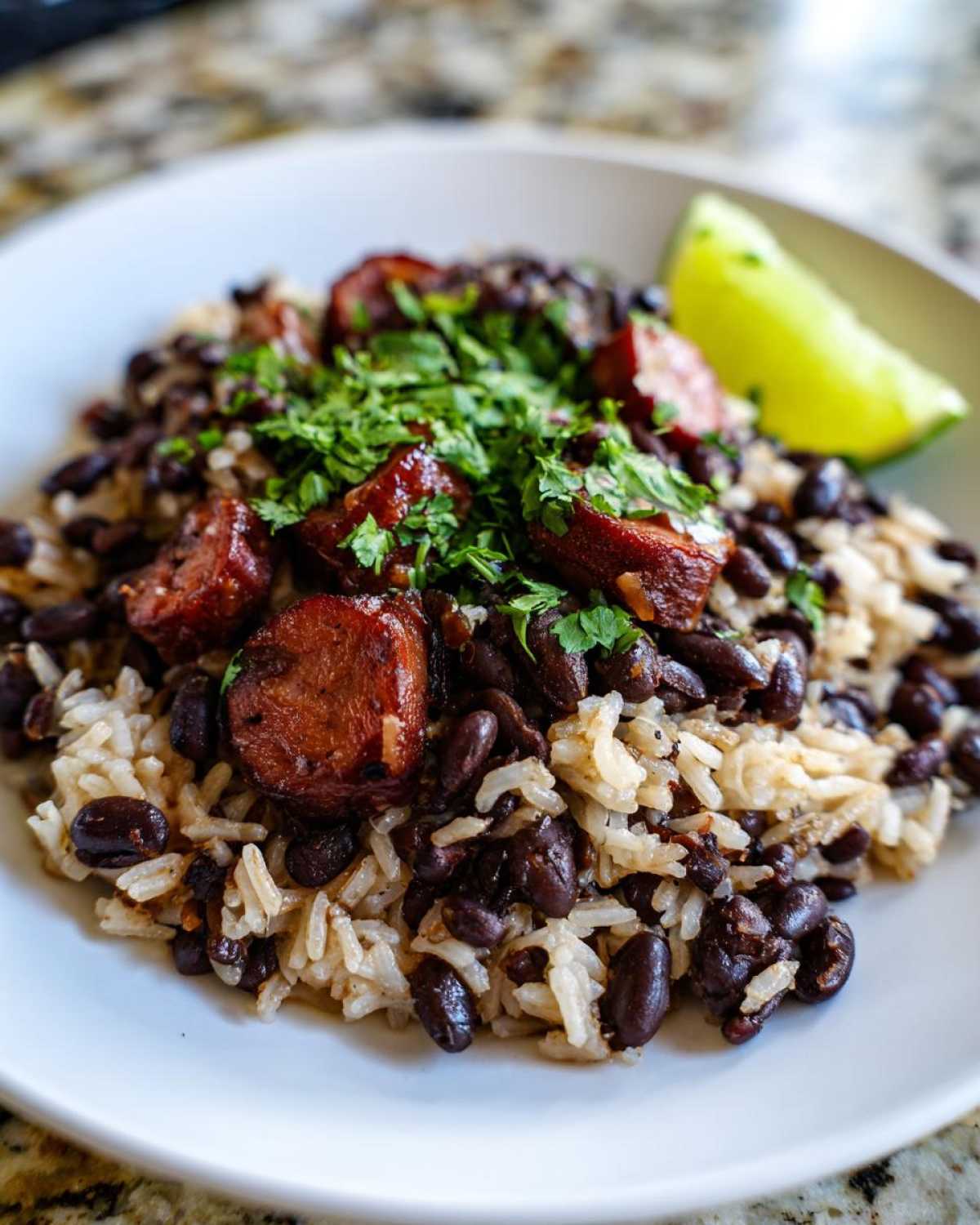 Close-up of savory black beans and rice mixed with sliced sausage, topped with cilantro, served with a lime wedge.