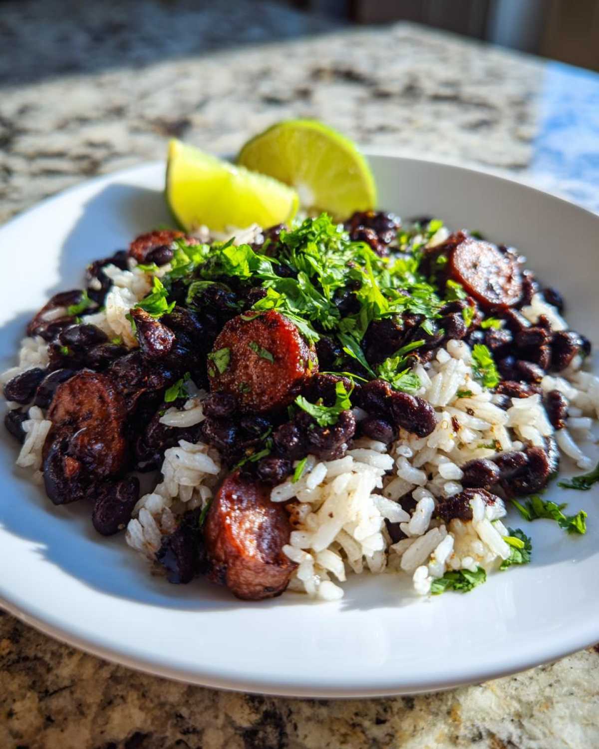 A close-up of savory black beans and rice mixed with sliced sausage, topped with fresh cilantro and lime wedges.