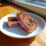 Two perfectly seared tuna steak halves showing a bright pink, rare center, seasoned with pepper.