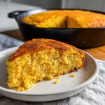 A close-up of a golden slice of skillet cornbread served on a white plate, with the rest of the cornbread visible in the cast iron skillet behind it.