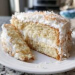 A close-up of a moist slice of coconut cake layered with white frosting and topped with toasted coconut flakes.