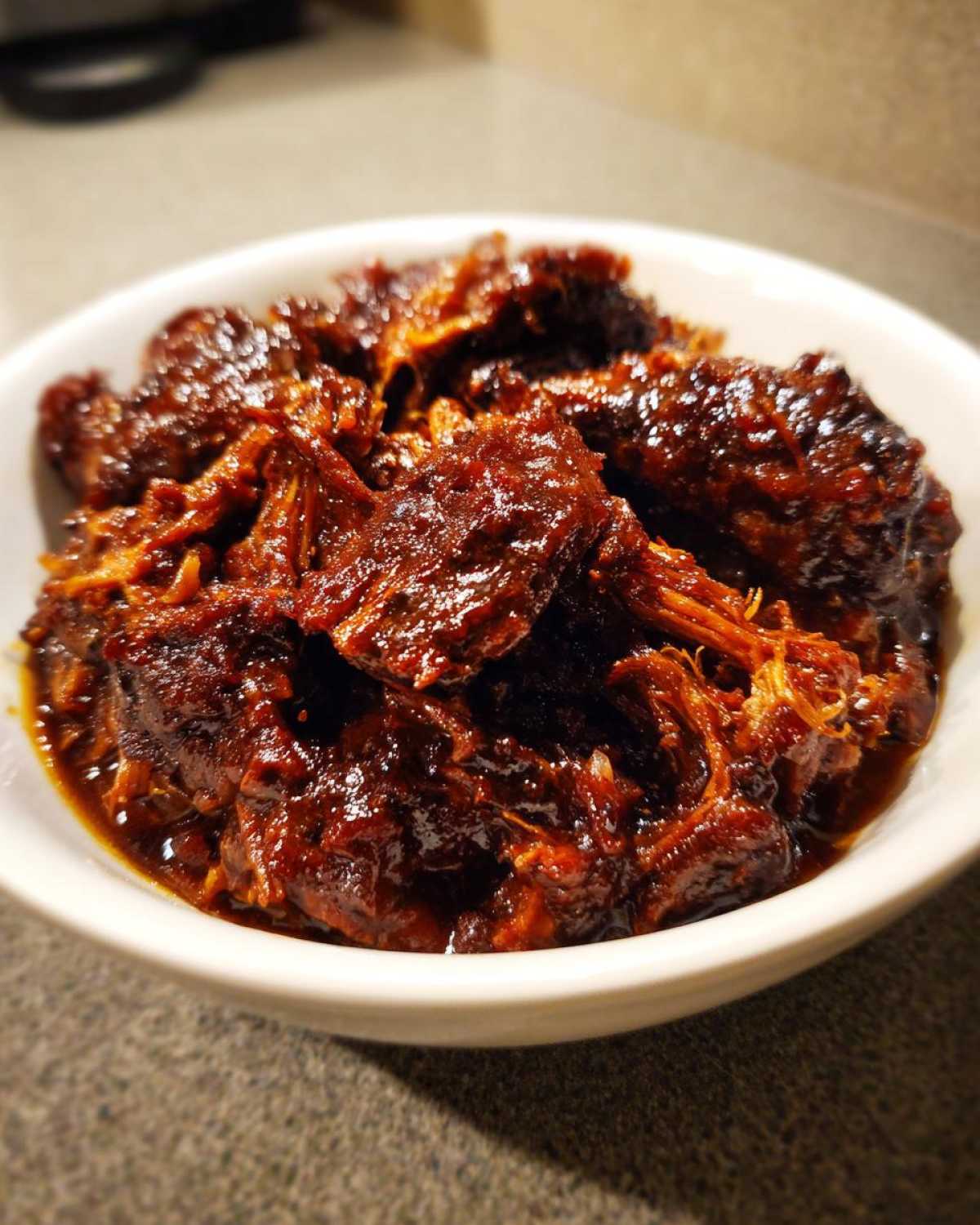 A white bowl filled with shredded, saucy Slow Cooker Pulled Pork glistening under the light.