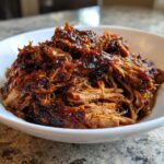 Close-up of rich, saucy Slow Cooker Pulled Pork shredded and piled high in a white bowl.