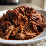 A close-up of tender, shredded Slow Cooker Pulled Pork coated in rich barbecue sauce piled high on a white plate.