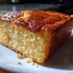 Close-up of a moist, golden slice of sourdough cornbread with a shiny, caramelized top served on a white plate.