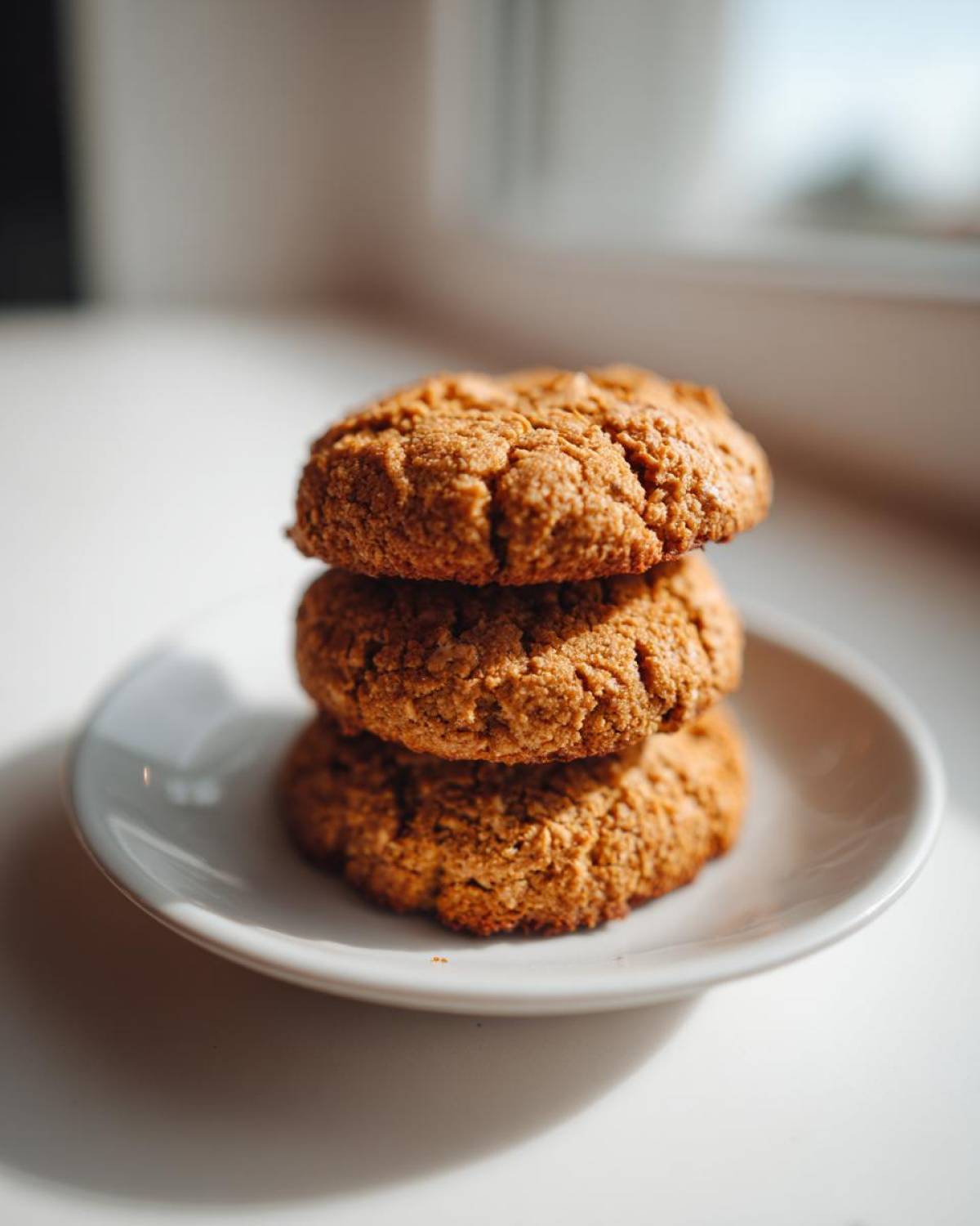 Three textured, golden-brown breakfast cookie treats stacked on a small white plate near a window.