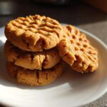 A stack of three golden brown peanut butter cookies with classic fork crosshatch marks, resting on a white plate.