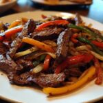 Close-up of perfectly cooked steak fajitas with colorful strips of red, yellow, and green peppers and caramelized onions on a white plate.