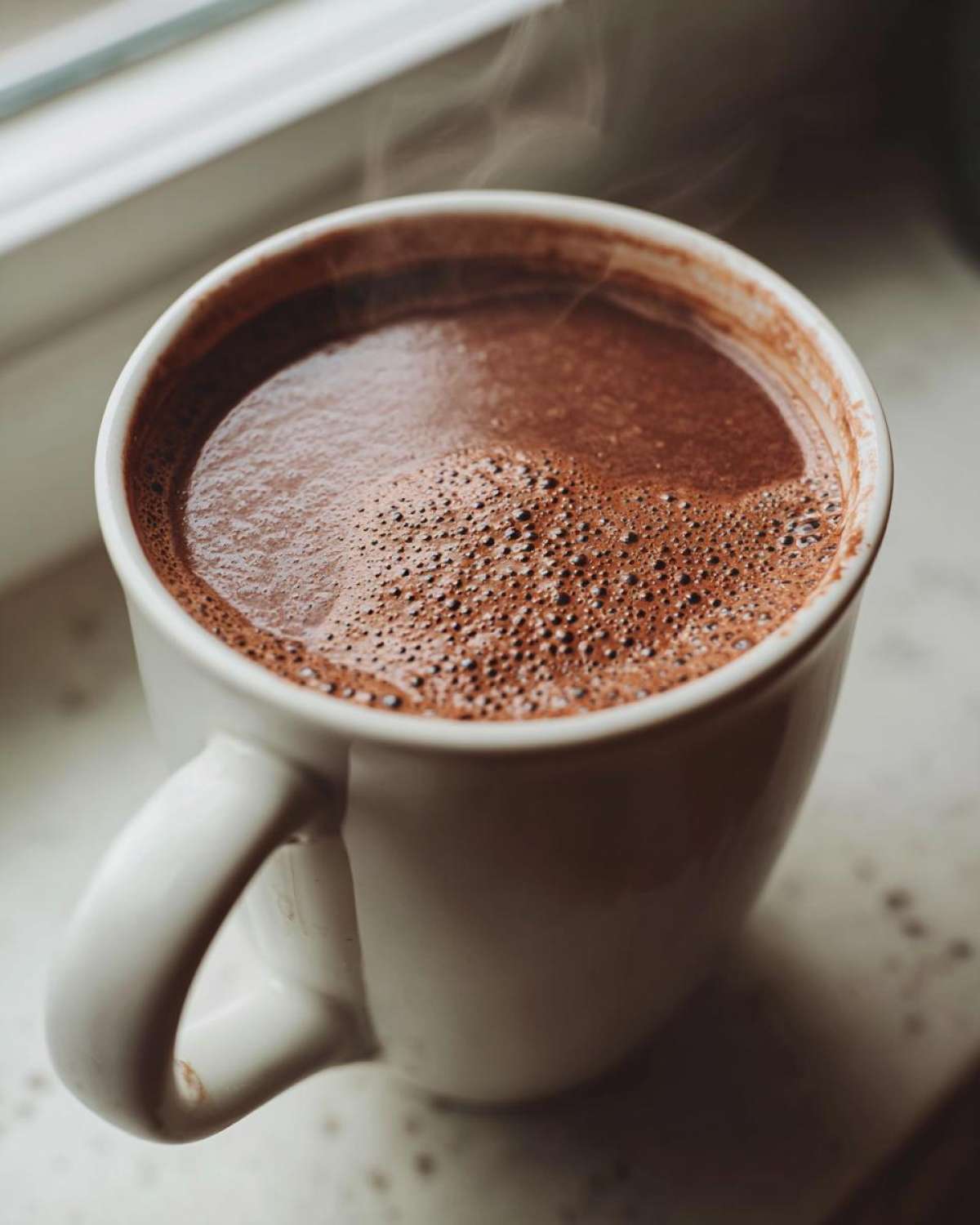 Close-up of a white mug filled with steaming, rich brown hot cocoa mix resting near a window.