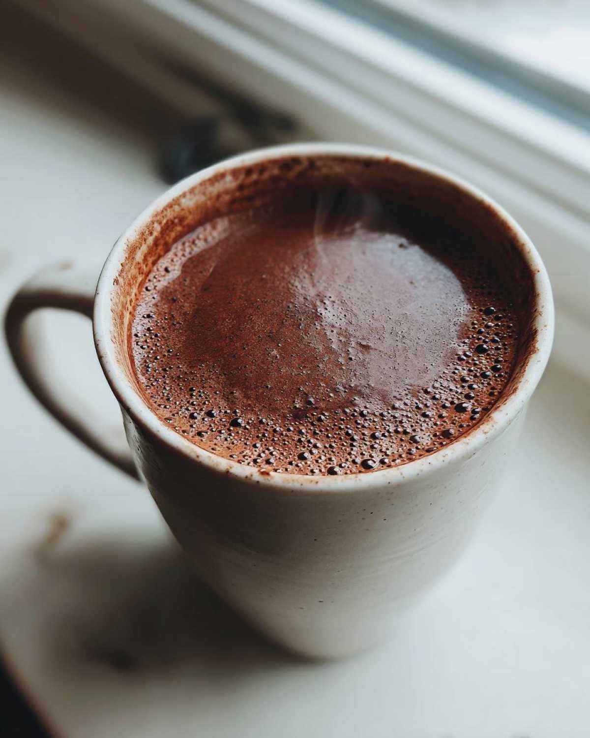 Close-up of a steaming mug filled with rich, dark brown hot cocoa mix, resting on a white windowsill.