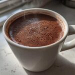 Close-up of a steaming white mug filled with rich, dark brown hot cocoa mix, bubbling slightly.