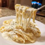 A fork lifts fettuccine noodles coated in creamy olive garden alfredo sauce, showing a dramatic cheese pull.