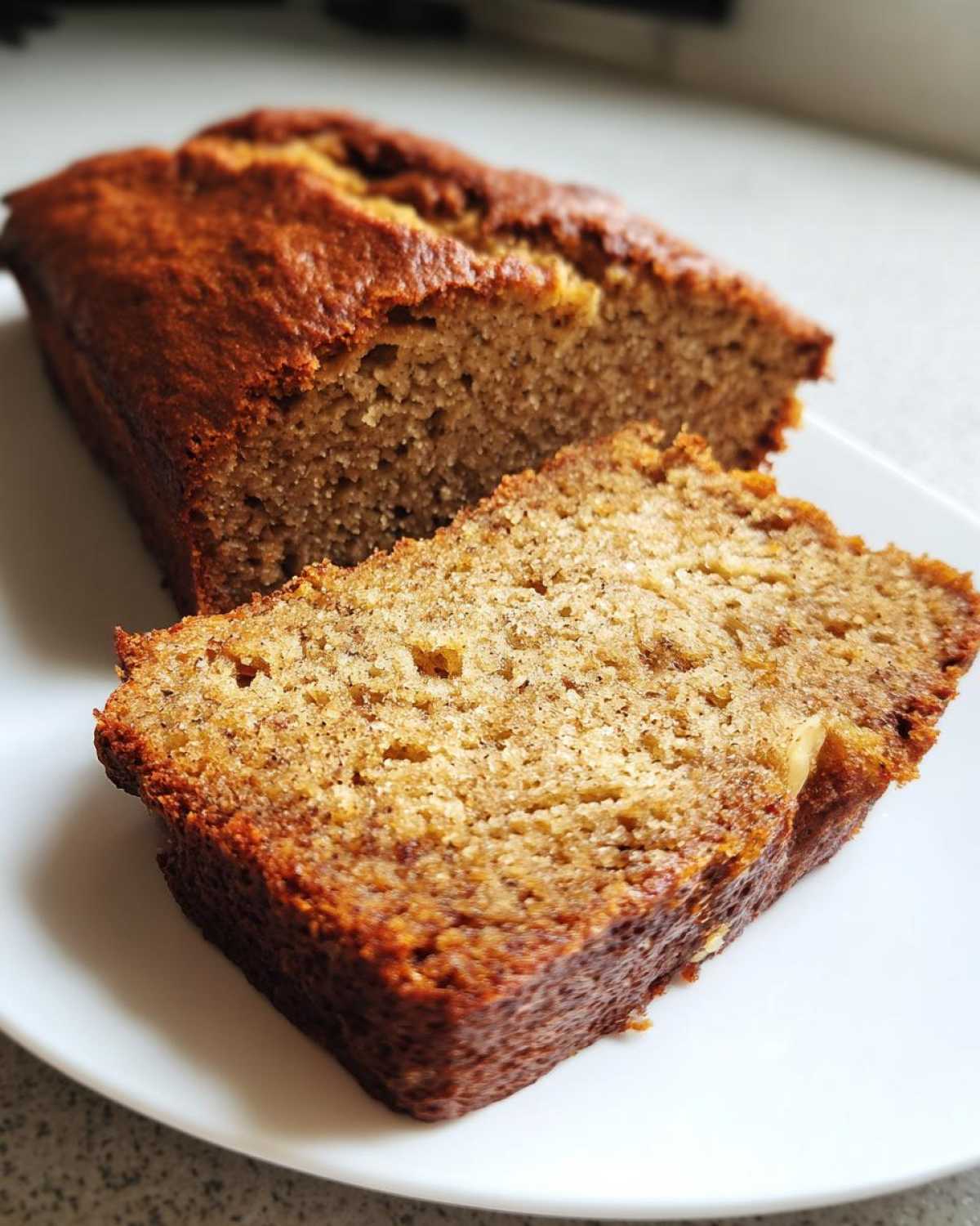 Close-up of a slice of moist sugar free banana bread next to the loaf on a white plate.