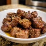 Close-up of tender, bite-sized pieces of crockpot steak coated in a savory garlic butter sauce in a white bowl.