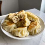 A white plate piled high with golden-brown, pan-fried Traditional Chinese Dumplings, one cut open to show the savory filling.