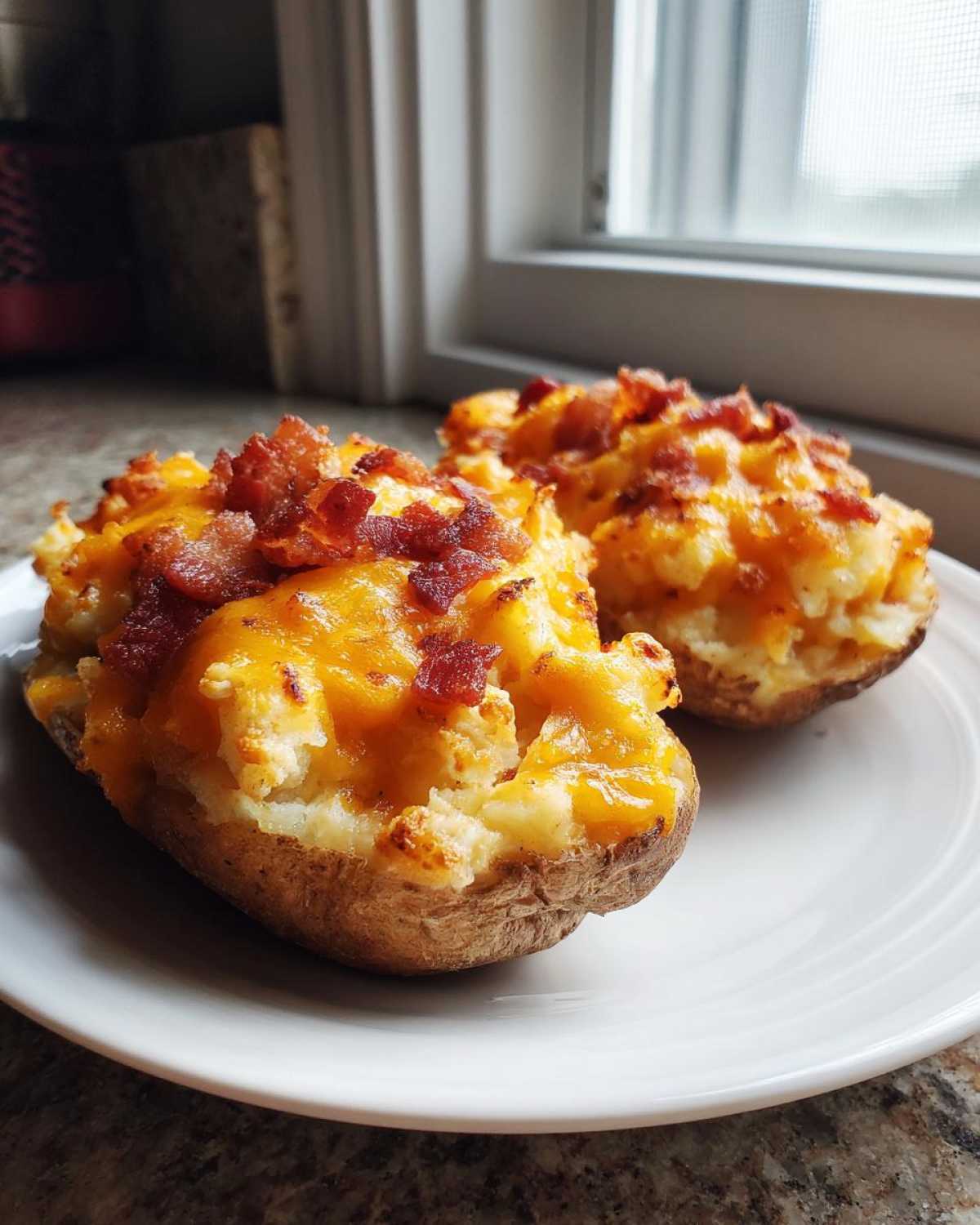 Two perfectly prepared twice baked potatoes loaded with melted cheddar cheese and crispy bacon bits on a white plate.