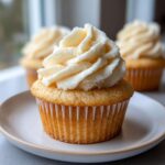 Close-up of a moist vanilla cupcake with a tall swirl of creamy white vanilla frosting, sitting on a small plate.
