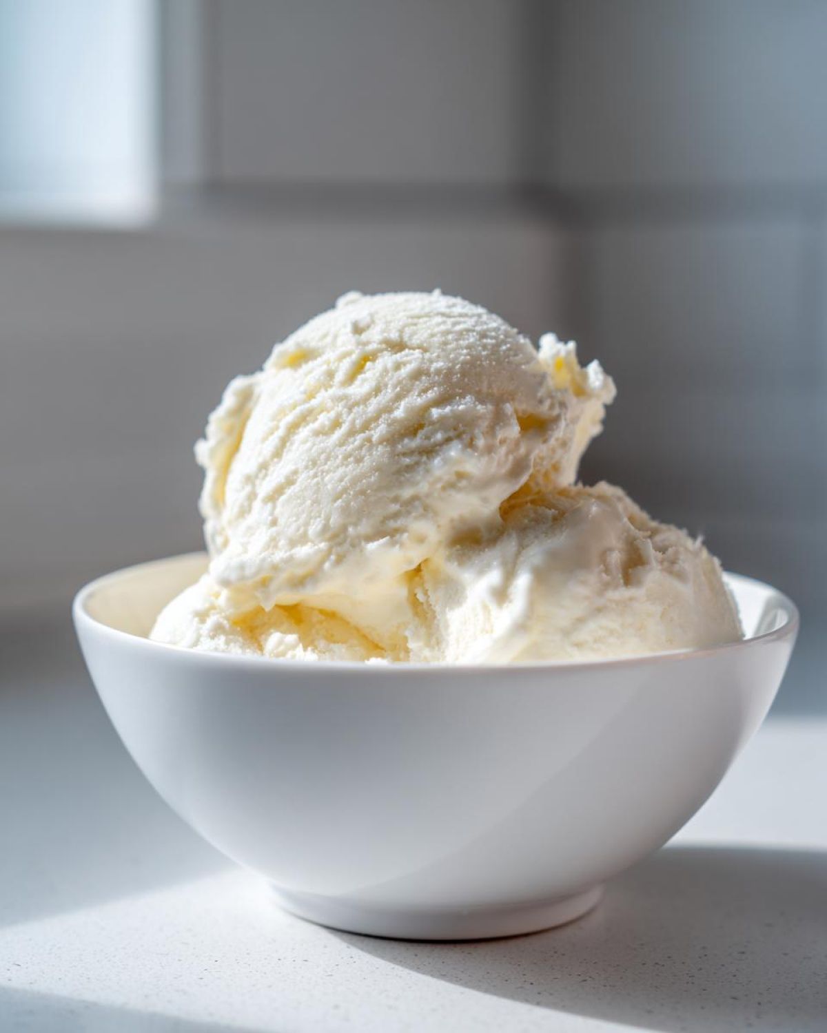 Two scoops of creamy vanilla protein ice cream served in a simple white bowl on a light countertop.