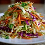 A mound of fresh cabbage salad featuring shredded white cabbage, purple cabbage, bright orange carrots, and green parsley on a white plate.