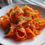 Close-up of a vibrant raw carrot salad made with shaved carrot ribbons, topped with sesame seeds and green onions.
