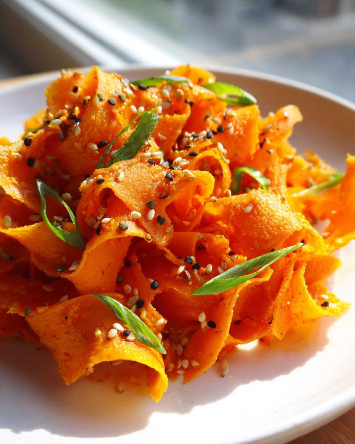 Close-up of bright orange shaved ribbons in a raw carrot salad, topped with black and white sesame seeds and green onions.