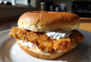 Close-up of a golden brown, crispy Air Fryer Crispy Fish Sandwich topped with white tartar sauce on a brioche bun.