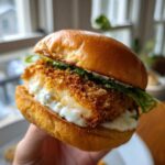 A hand holding a perfectly assembled Air Fryer Crispy Fish Sandwich featuring a golden, breaded fillet and creamy tartar sauce.