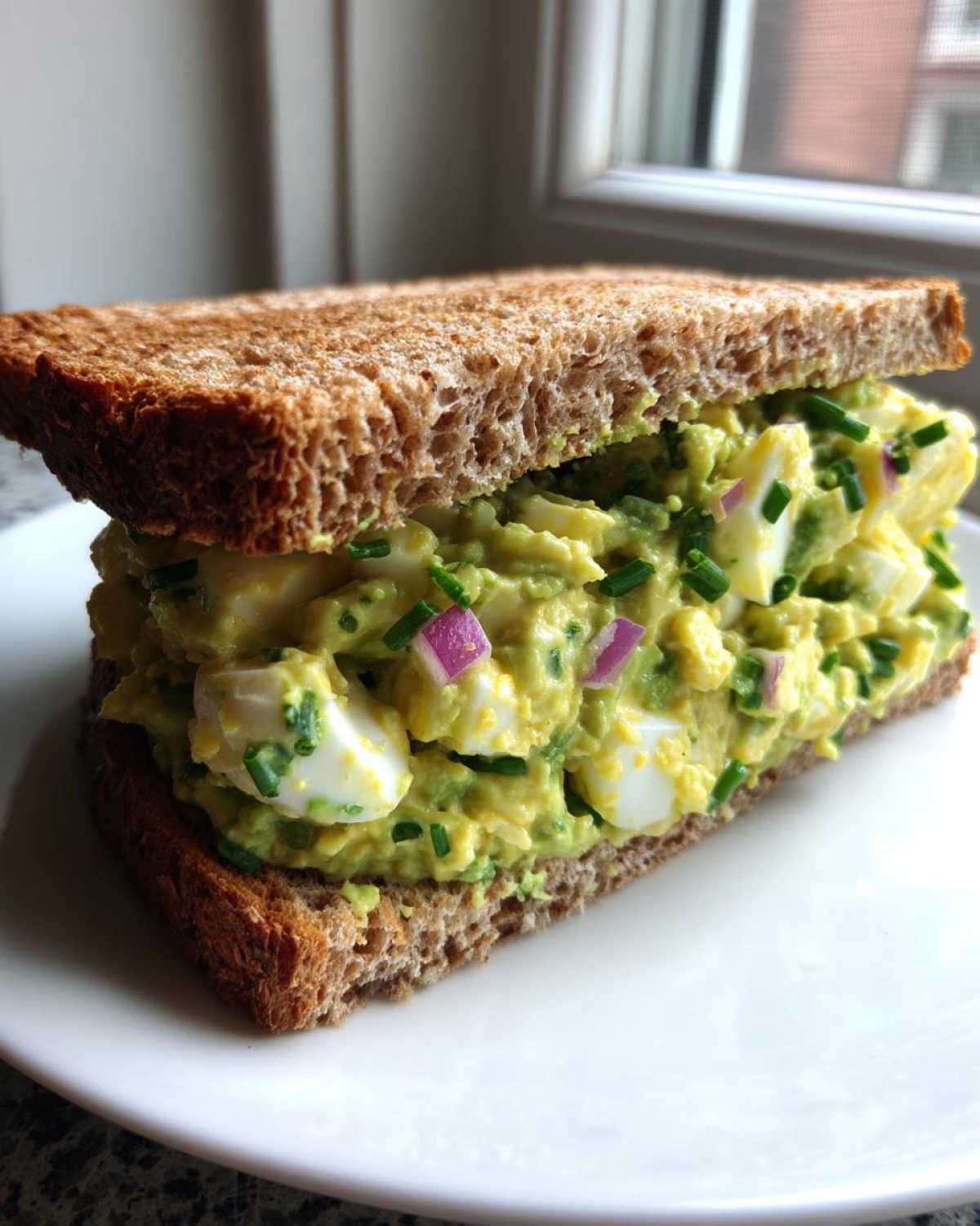 A thick, loaded Avocado Egg Salad Sandwich made with whole wheat bread, showing chunks of egg and green avocado mash with red onion and chives.