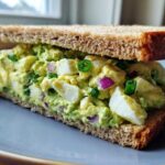 A thick-cut Avocado Egg Salad Sandwich made with whole wheat bread, showing chunks of egg, green avocado mash, red onion, and chives.