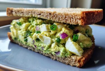 A thick-cut Avocado Egg Salad Sandwich made with whole wheat bread, showing chunks of egg, green avocado mash, red onion, and chives.