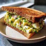 A hearty Avocado Egg Salad Sandwich made with chunks of hard-boiled egg, avocado mash, red onion, and chives, served on toasted whole wheat bread.